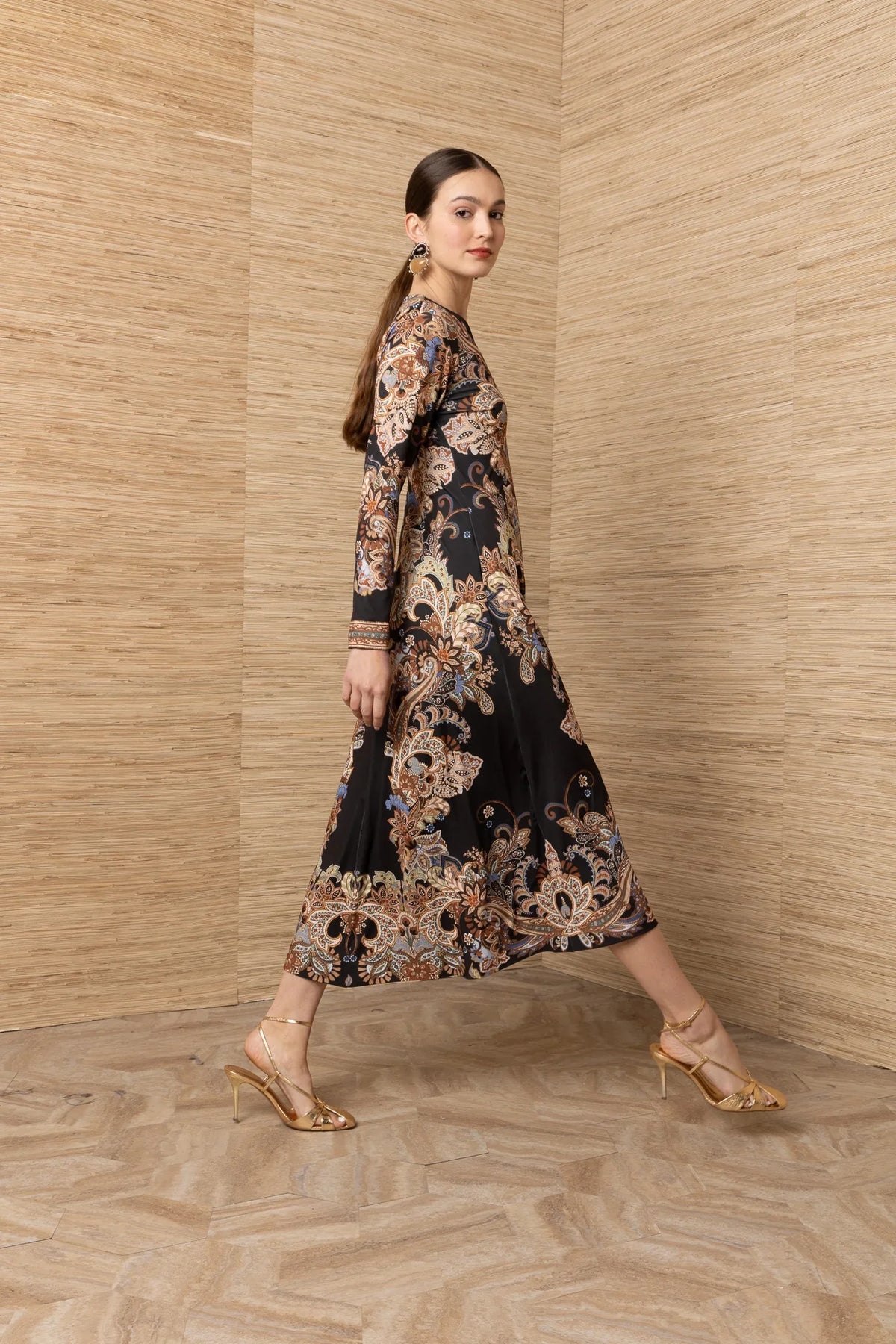 Woman in a black long-sleeve midi dress with gold and blue baroque pattern and gold heels.