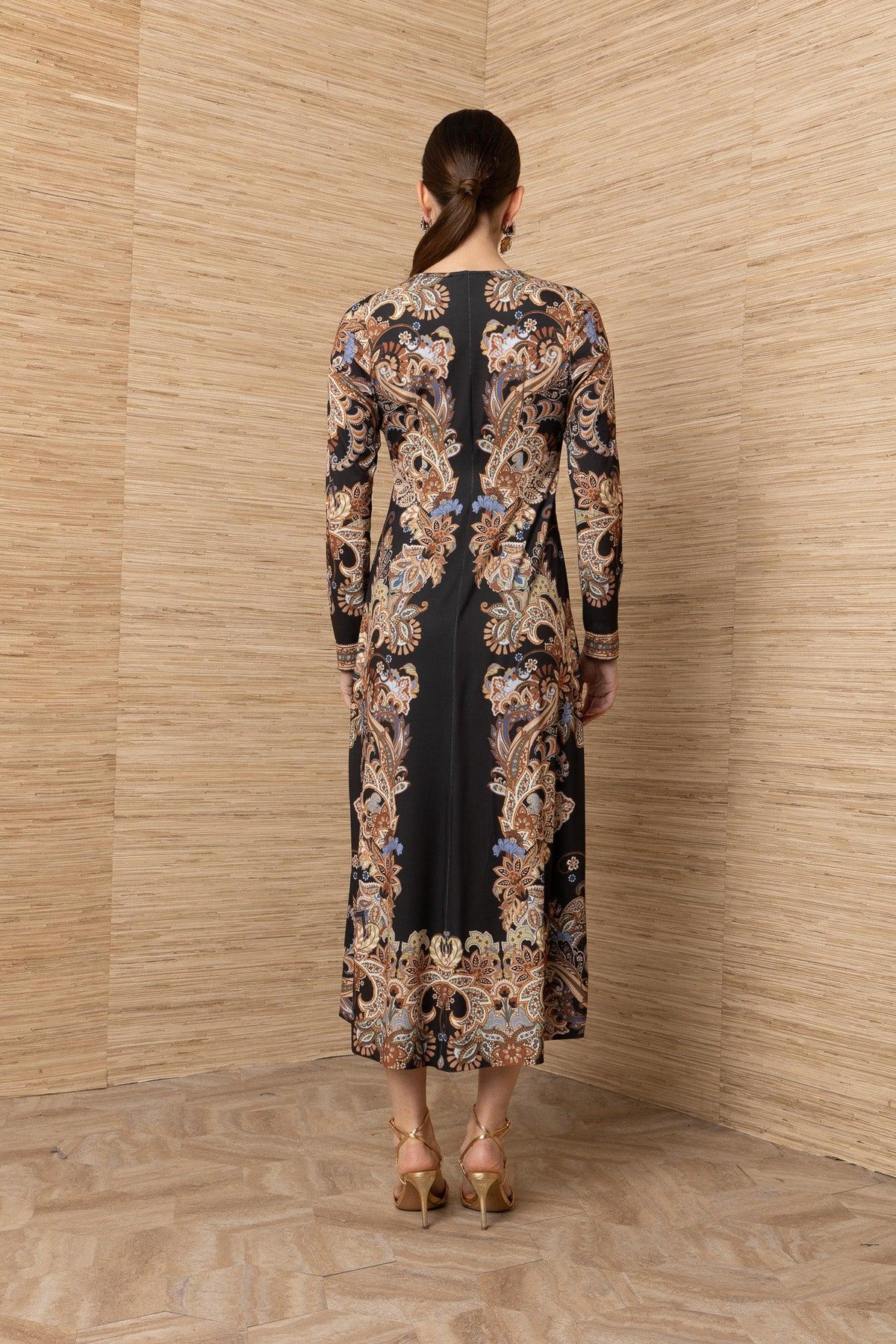 Back view of a woman wearing a long-sleeved black maxi dress with an ornate gold baroque print.
