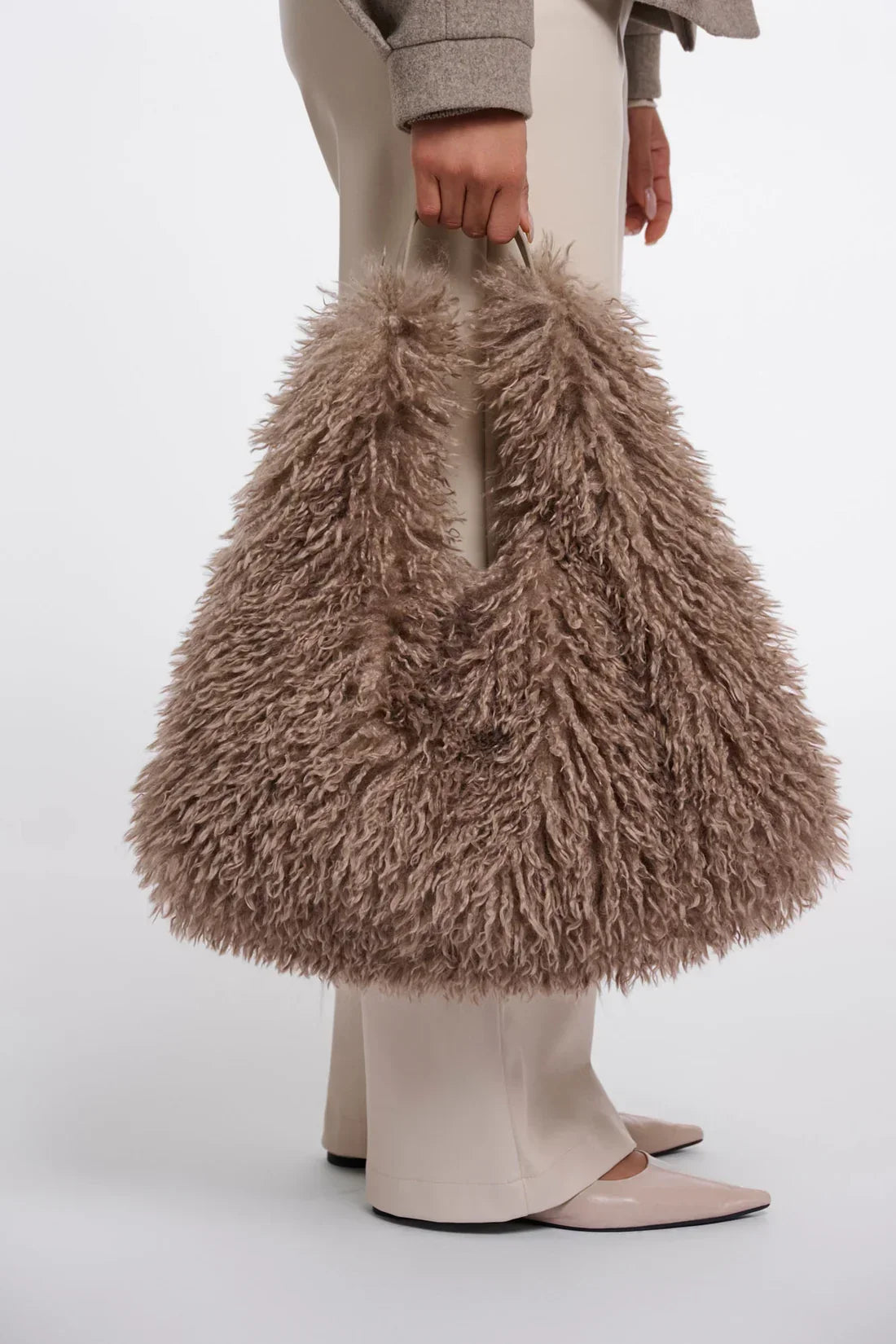 Woman holding a large, fluffy taupe faux fur handbag paired with cream trousers.