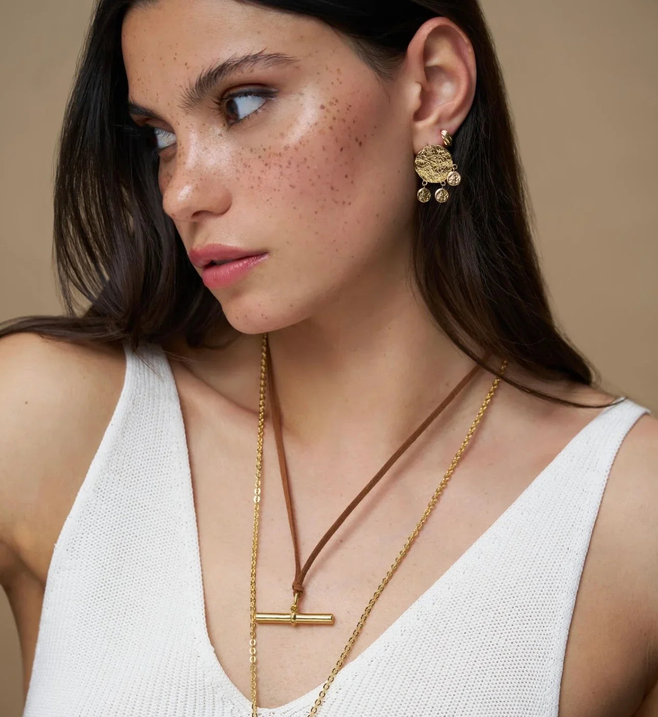 Woman wearing gold drop earrings and layered gold necklaces with a brown cord pendant.