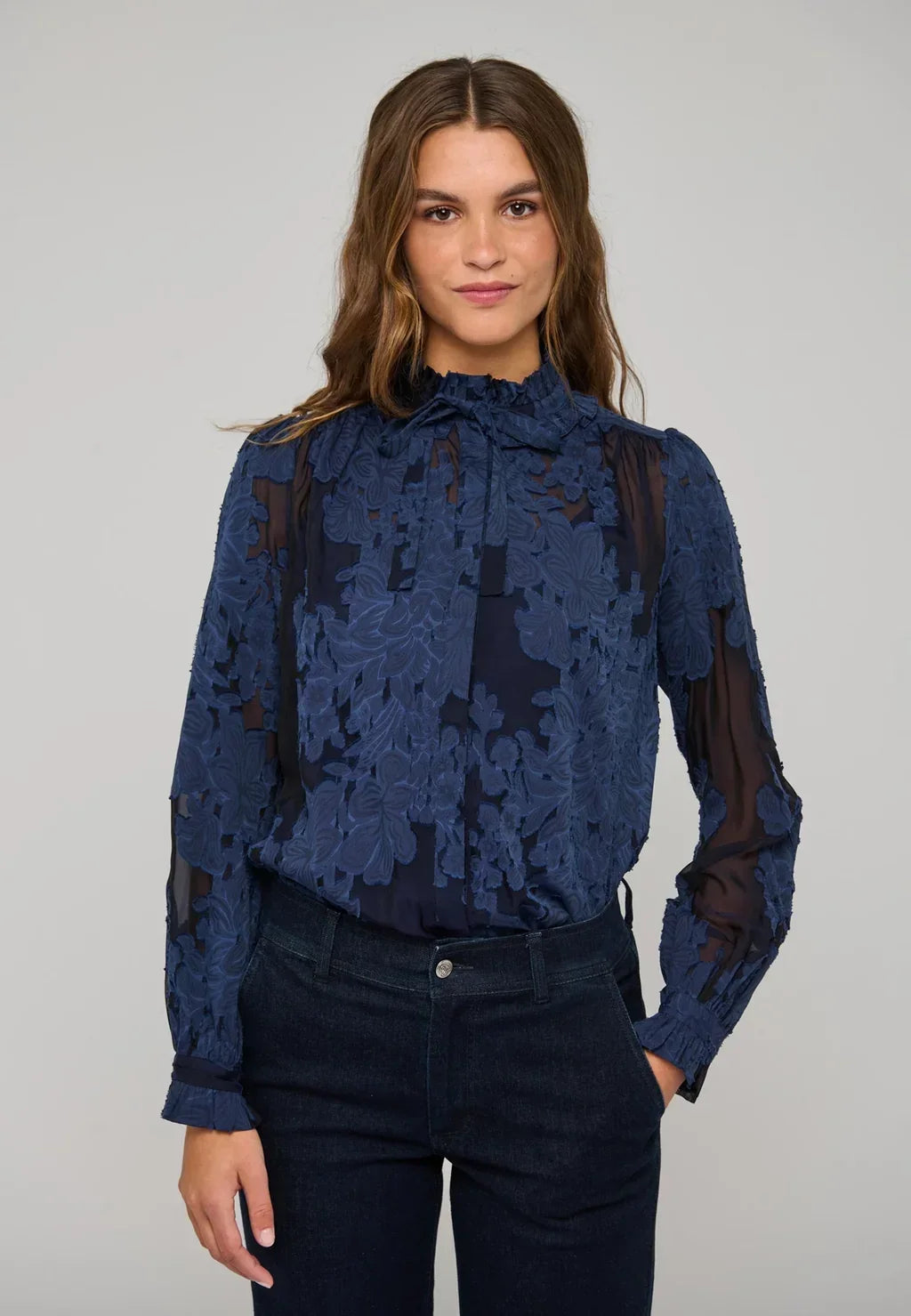 Woman wearing a navy blue floral lace long-sleeved blouse and dark denim jeans.