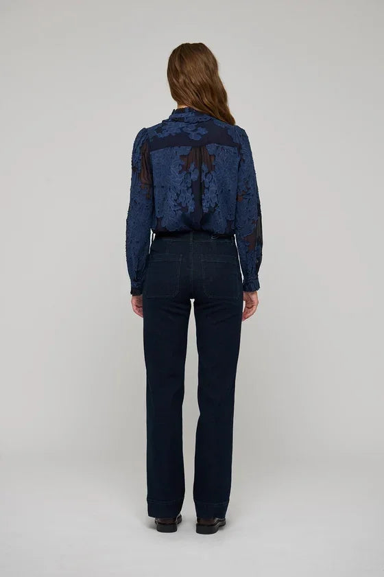 Back view of a woman wearing a blue floral lace blouse and dark navy wide-leg trousers.