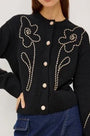 Woman wearing a black knit cardigan with white floral rope embroidery and cream buttons.