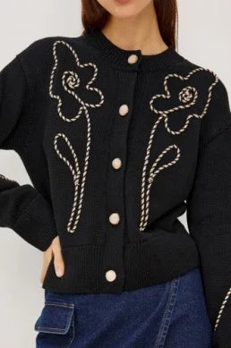 Woman wearing a black knit cardigan with white floral rope embroidery and cream buttons.