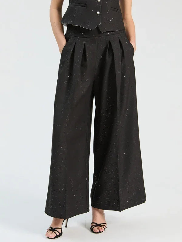 Woman wearing black wide-leg pleated trousers with subtle shimmer and a black vest.